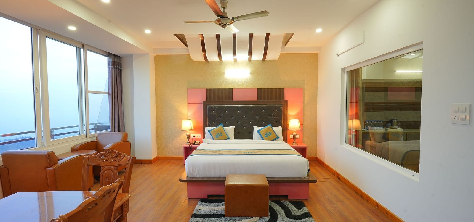 Shot of the suite room featuring a king size bed, wooden floor, carpet, a separate seating area and an expansive window |Shot o deluxe room eaturing king size bed, bed side lamps, two couches and a window | DLS Hotel Shiva Sanctuary and Spa